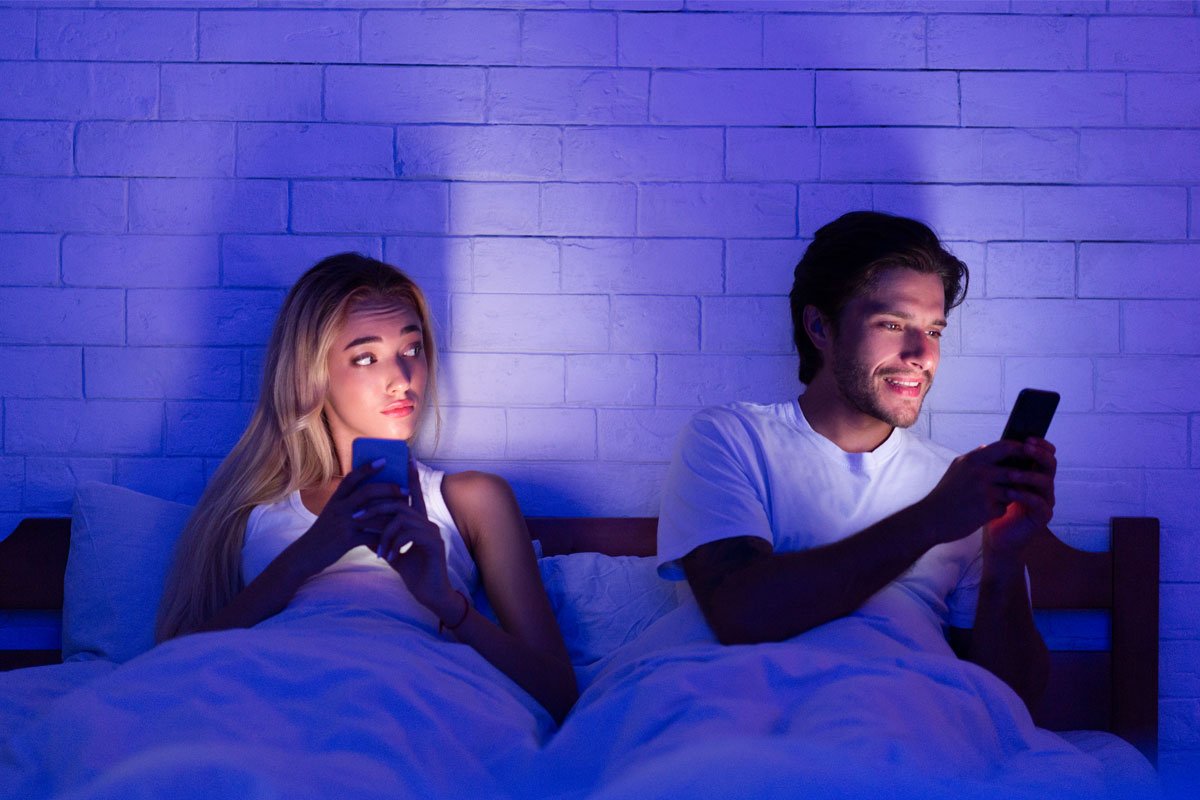 A young couple in bed at night, each looking at their phones and seemingly ignoring each other. | Forensic Discovery