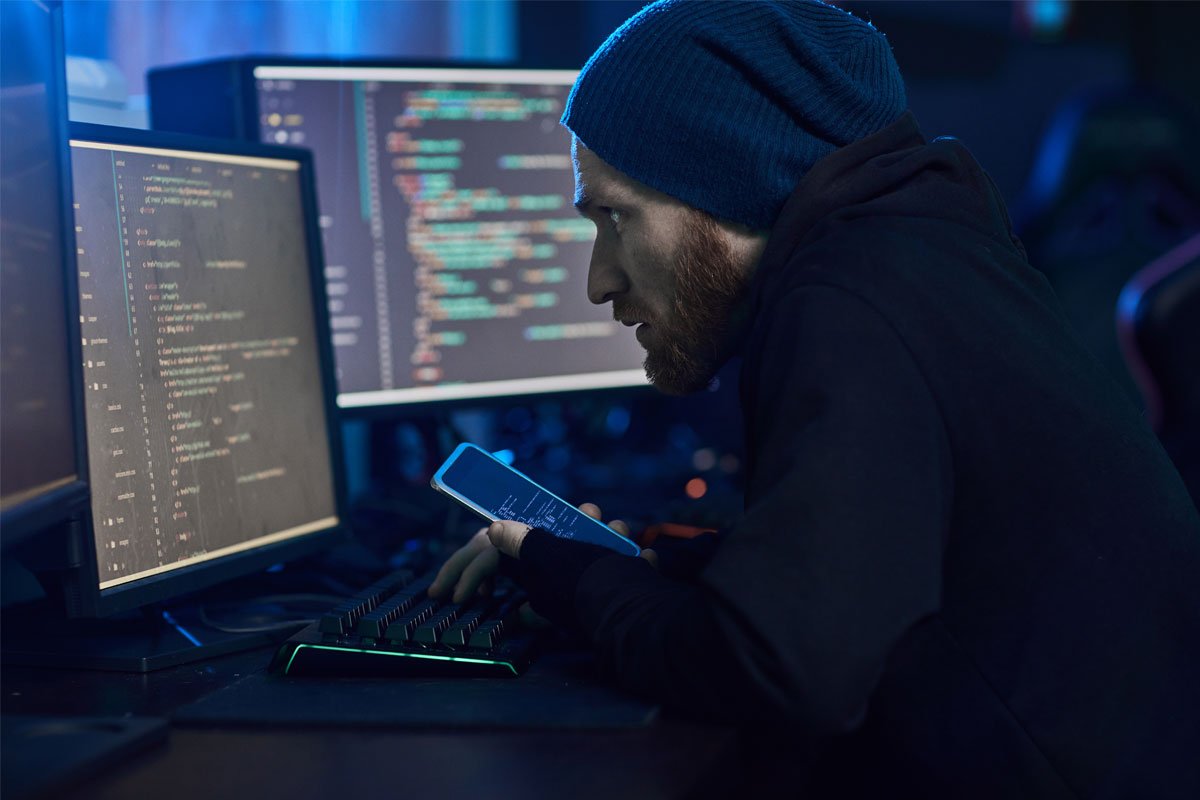 Person in a blue beanie looking at a phone while working on a computer with lines of code visible on the screen. | Forensic Discovery