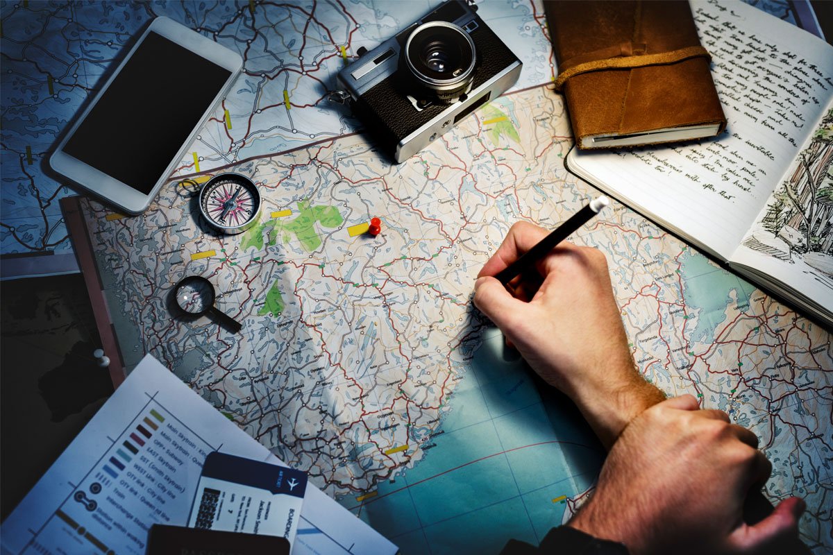 A person plans a trip, tracing a route on a map with a pencil. Surrounding the map are a compass, magnifying glass, smartphone, camera, journal, and travel documents. | Forensic Discovery