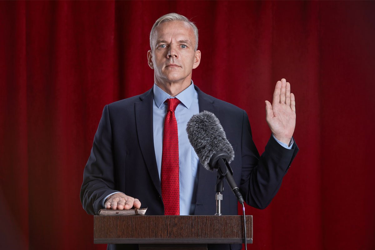 A man in a suit raises his right hand in front of a red curtain, appearing to take an oath at a podium with a microphone. | Forensic Discovery